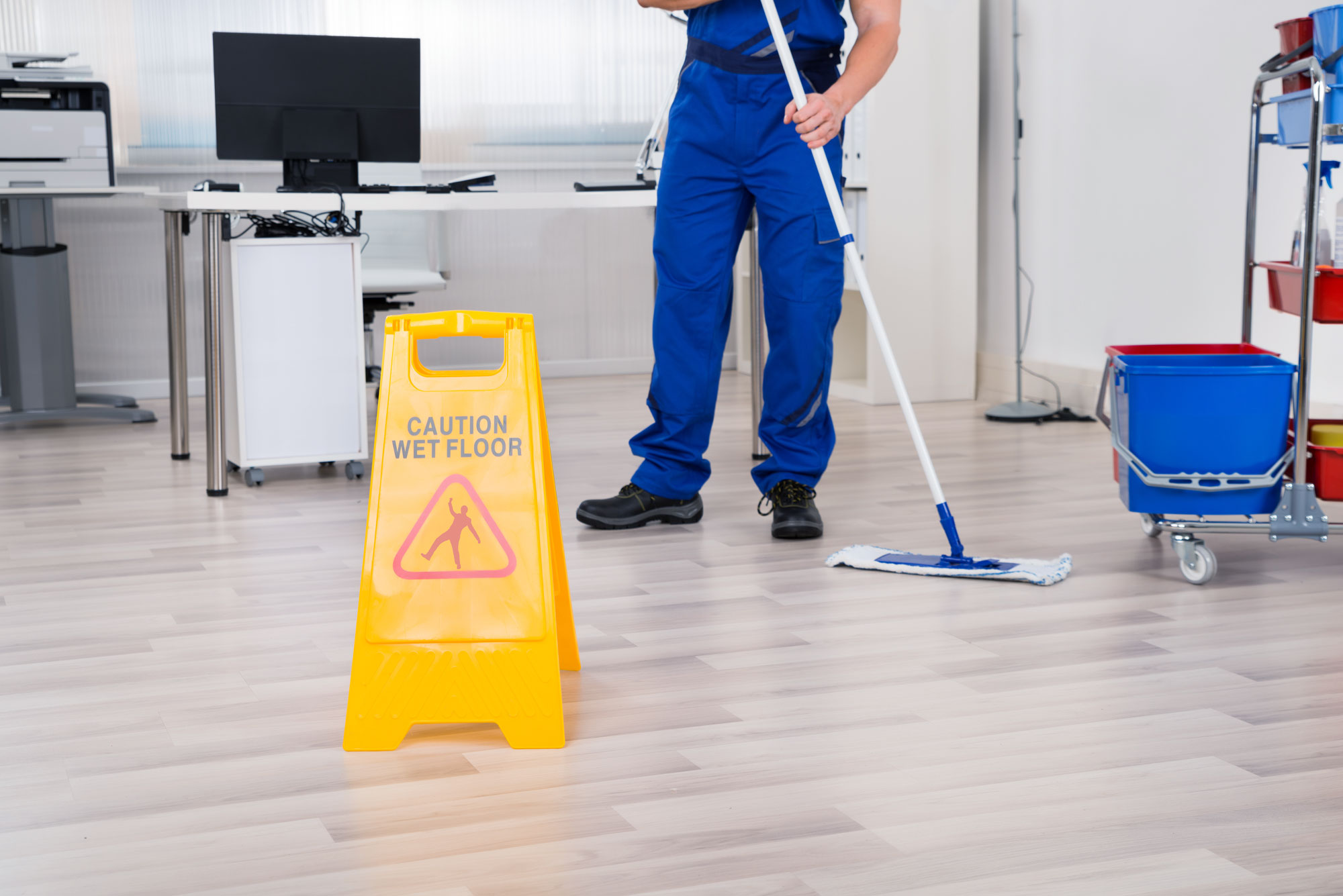 janitor wearing blue uniform mopping commercial office with wet floor sign propped up