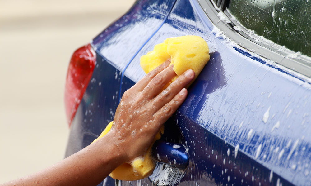 hand with yellow sponge washing side of blue car